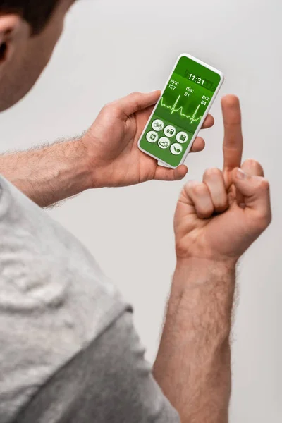 Cropped view of man using smartphone with health app and showing middle finger, isolated on grey — Stock Photo