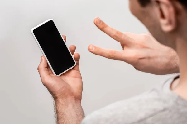 Обрізаний знак людини, що показує знак перемоги, тримаючи смартфон з порожнім екраном ізольовано на сірому — стокове фото