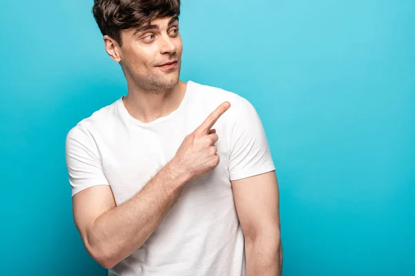 Joven guapo sonriendo mientras mira hacia otro lado y señala con el dedo sobre el fondo azul — Stock Photo