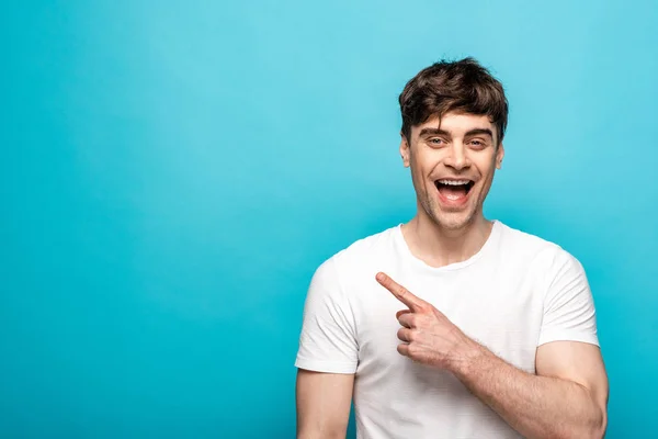 Fröhlicher junger Mann zeigt mit dem Finger und blickt in die Kamera auf blauem Hintergrund — Stockfoto