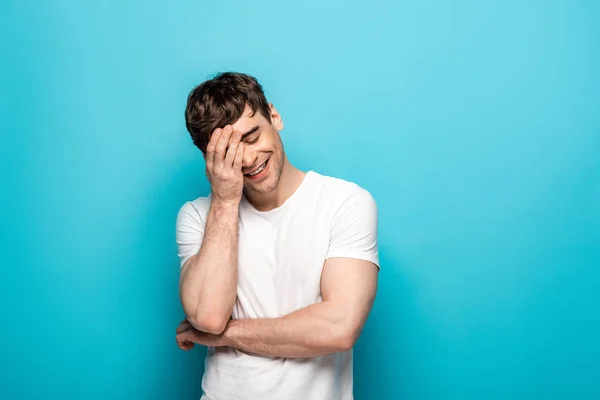 Heureux jeune homme souriant tout en tenant la main sur le visage sur fond bleu — Photo de stock