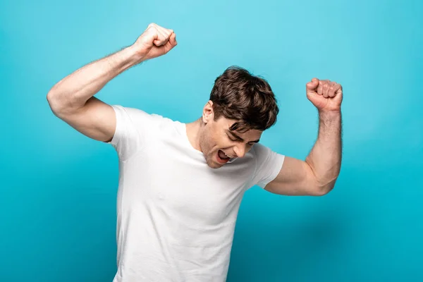 Happy young man showing winner gesture with closed eyes on blue background — Stock Photo