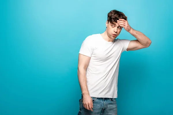 Choqué jeune homme regardant loin tout en tenant la main sur la tête sur fond bleu — Photo de stock