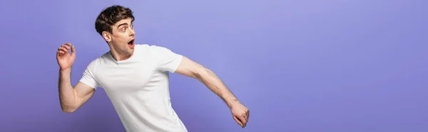 Panoramic shot of scared young man running away and looking back isolated on blue — Stock Photo