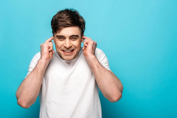 Jeune homme positif bouchant les oreilles avec les doigts sur fond bleu — Photo de stock