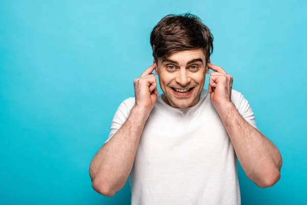 Fröhlicher junger Mann, der mit den Fingern Ohren stopft, während er in die Kamera auf blauem Hintergrund schaut — Stockfoto