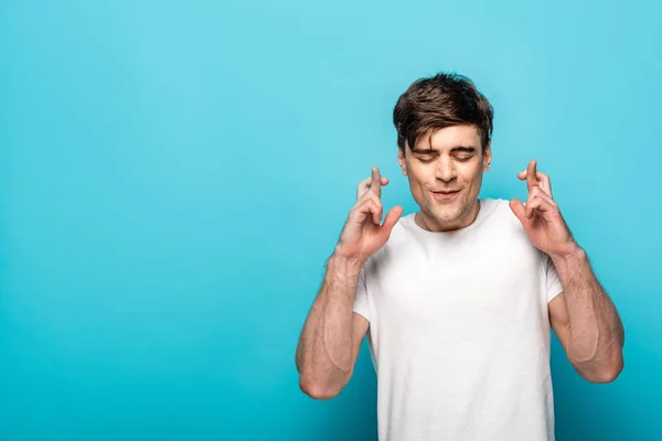 Jeune homme positif tenant les doigts croisés avec les yeux fermés sur fond bleu — Photo de stock