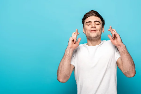 Positive young man holding crossed fingers with closed eyes on blue background — Stock Photo