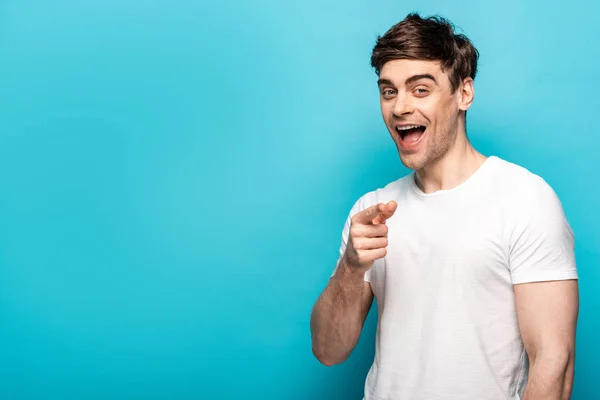 Fröhlicher junger Mann zeigt mit dem Finger und lächelt in die Kamera auf blauem Hintergrund — Stockfoto