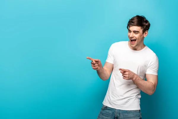 Cheerful young man pointing with fingers and looking away on blue background — Stock Photo