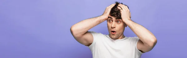 Panoramic shot of shocked young man holding hands on head and looking away on blue background — Stock Photo