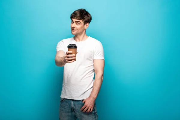 Souriant jeune homme tenant tasse en papier dans la main tendue et regardant la caméra sur fond bleu — Photo de stock