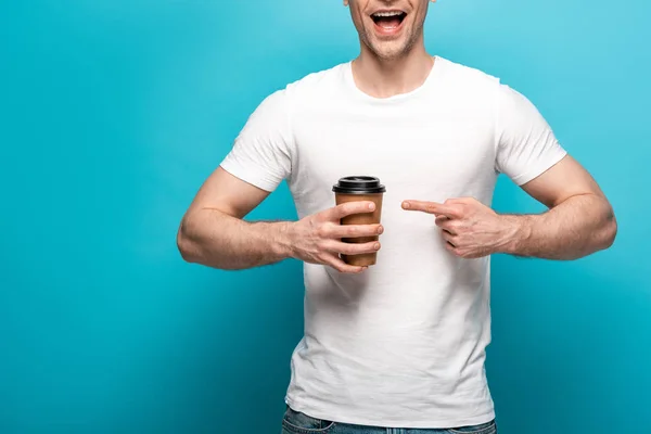 Обрезанный вид улыбающегося мужчины, указывающего пальцем на бумажную чашку на синем фоне — стоковое фото