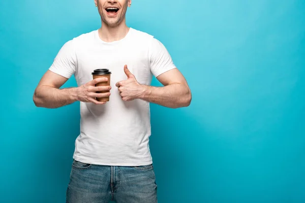Recortado vista de sonriente hombre sosteniendo café para ir y mostrando el pulgar hacia arriba sobre fondo azul - foto de stock