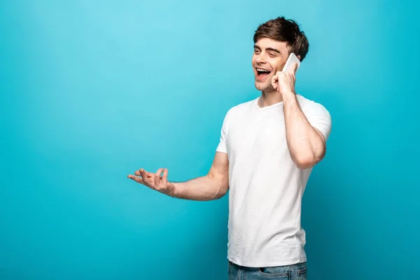 Веселый молодой человек жестикулирует, разговаривая на смартфоне и глядя в сторону на голубом фоне — стоковое фото