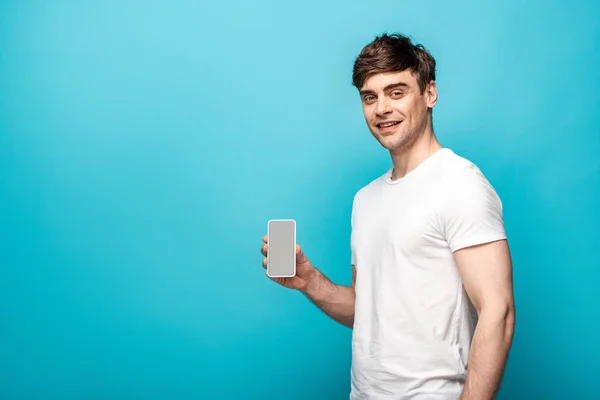 Joven guapo mostrando teléfono inteligente con pantalla en blanco y sonriendo a la cámara sobre fondo azul - foto de stock