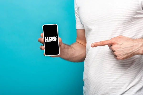 KYIV, UCRANIA - 16 DE MAYO DE 2019: Vista recortada del hombre en camiseta blanca apuntando con el dedo al teléfono inteligente con la aplicación HBO en pantalla sobre fondo azul . - foto de stock