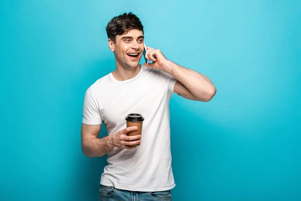 Joven sonriente hablando en el teléfono inteligente mientras sostiene el café para ir y mirar hacia otro lado en el fondo azul - foto de stock