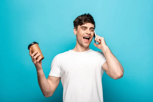 Cheerful young man talking on smartphone and holding coffee to go on blue background — Stock Photo