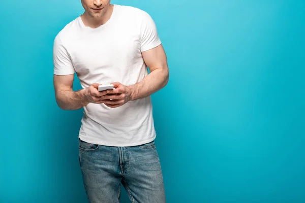 Cropped view of young man in white t-shirt and blue jeans using smartphone on blue background — Stock Photo