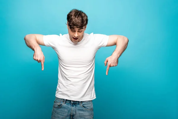 Jeune homme en t-shirt blanc pointant vers le bas avec les doigts sur fond bleu — Photo de stock