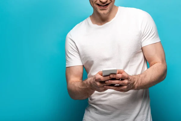 Vista recortada de un joven sonriente usando un teléfono inteligente sobre un fondo azul - foto de stock