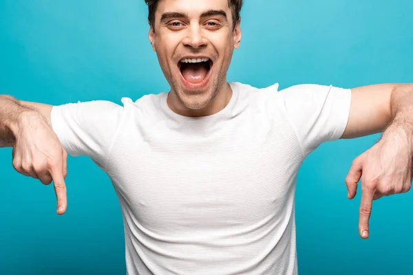 Joven alegre apuntando hacia abajo con los dedos y sonriendo a la cámara sobre fondo azul - foto de stock