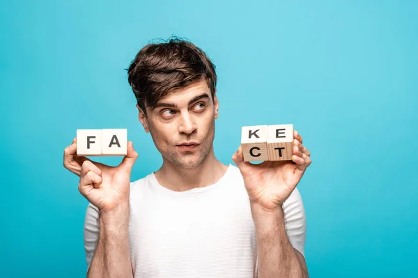 Thoughtful man looking away while holding wooden cubes with fake fact lettering on blue background — Stock Photo