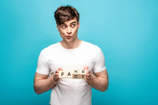 Desanimado jovem segurando cubos de madeira com falso fato lettering no fundo azul — Fotografia de Stock