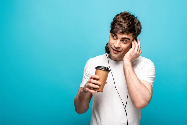 Cheerful young man in headphones holding coffee to go and looking away on blue background — Stock Photo