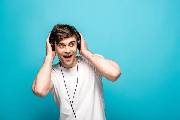 Sorrindo jovem ouvindo música em fones de ouvido e olhando para o fundo azul — Fotografia de Stock