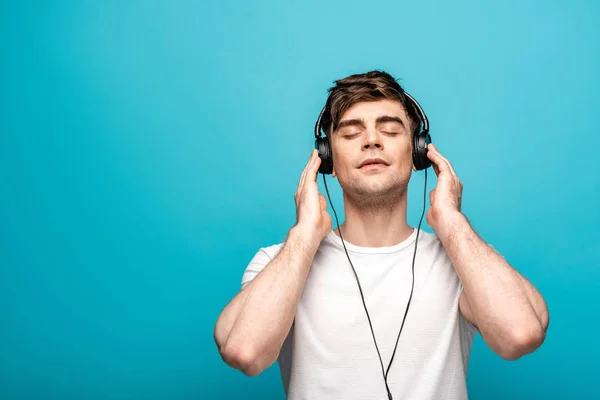 Jeune homme rêveur dans les écouteurs écouter de la musique avec les yeux fermés sur fond bleu — Photo de stock