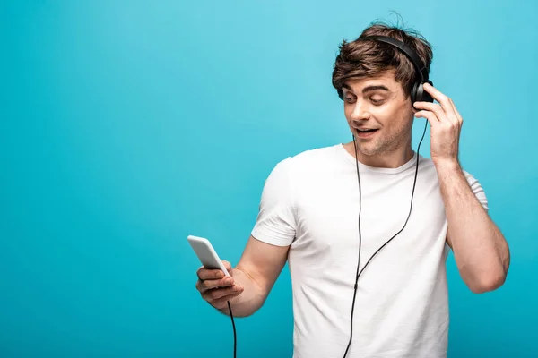 Handsome man listening music in headphones and using smartphone on blue background — Stock Photo