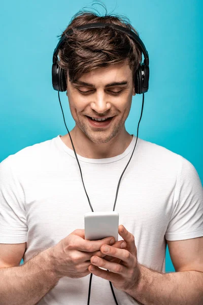 Cheerful young man listening music in headphones while using smartphone isolated on blue — Stock Photo