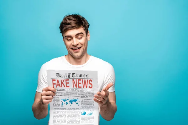 Улыбающийся молодой человек читает газету с фальшивыми новостями на синем фоне — стоковое фото