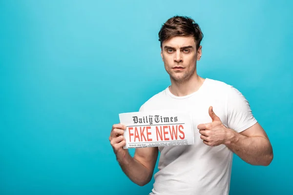 Joven serio mostrando el pulgar mientras sostiene el periódico con noticias falsas y mirando a la cámara en el fondo azul - foto de stock