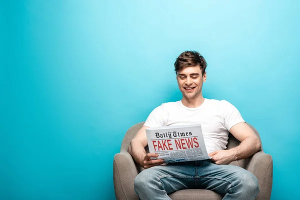 Fröhlicher junger Mann sitzt im Sessel und liest Fake News auf blauem Hintergrund — Stockfoto