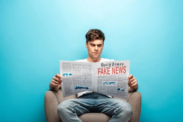 Wütender junger Mann liest Zeitung mit Fake News, während er im Sessel auf blauem Hintergrund sitzt — Stockfoto