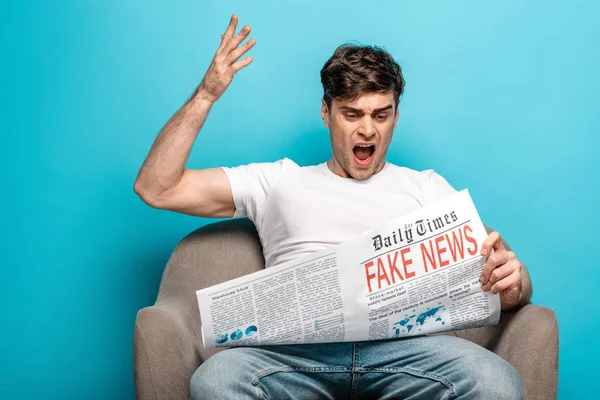 Hombre enojado haciendo gestos mientras está sentado en el sillón y leyendo el periódico con noticias falsas sobre fondo azul - foto de stock