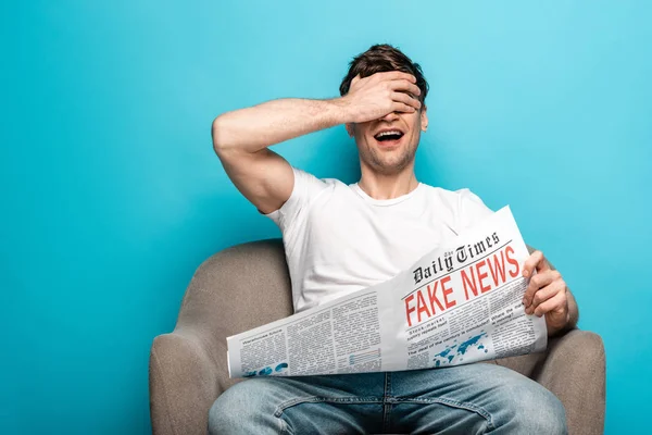 Jovem rindo enquanto sentado em poltrona e segurando jornal com notícias falsas sobre fundo azul — Fotografia de Stock
