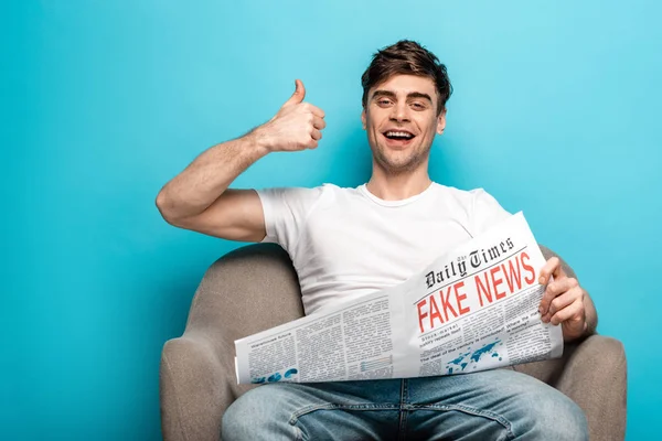 Fröhlicher junger Mann zeigt Daumen hoch, während er im Sessel sitzt und Zeitung mit Fake News auf blauem Hintergrund hält — Stockfoto