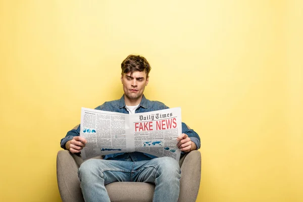 Serious young man sitting in armchair and reading newspaper with fake news on yellow background — Stock Photo