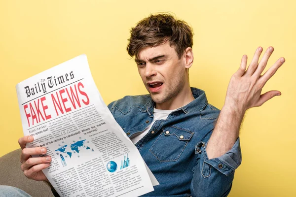 Unzufriedener junger Mann sitzt im Sessel und liest Fake News auf gelbem Hintergrund — Stockfoto