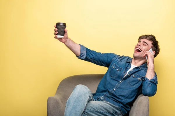 Joven emocionado hoding taza de papel mientras está sentado en el sillón y hablando en el teléfono inteligente sobre fondo amarillo - foto de stock