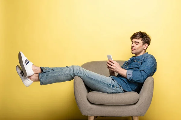 Handsome young man sitting in armchair and using smartphone on yellow background — Stock Photo