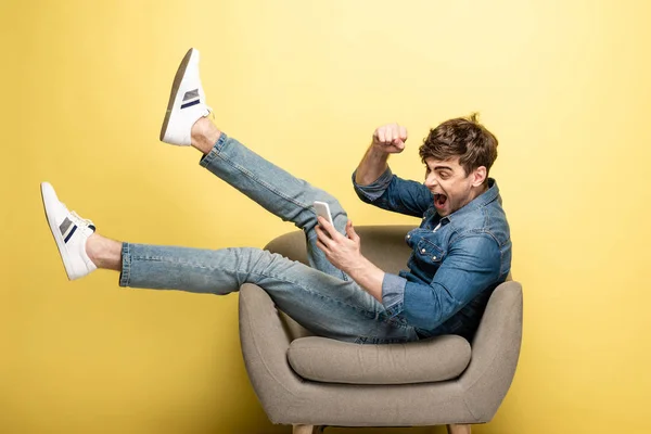 Excited young man showing yes gesture while sitting in armchair and using smartphone on yellow background — Stock Photo