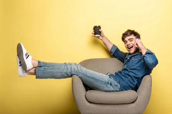 Hombre feliz sentado en sillón mientras habla en el teléfono inteligente y sostiene la taza de papel en el fondo amarillo - foto de stock