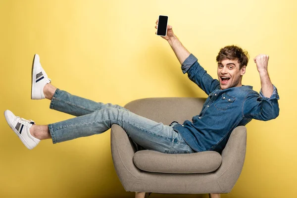 Joven emocionado mostrando gesto ganador mientras se sienta en el sillón y sostiene el teléfono inteligente con pantalla en blanco sobre fondo amarillo - foto de stock