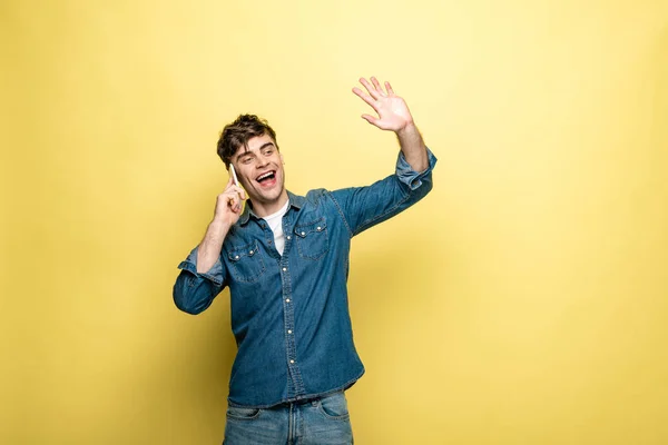 Fröhlicher junger Mann winkt mit der Hand und schaut weg, während er auf gelbem Hintergrund mit dem Smartphone spricht — Stockfoto