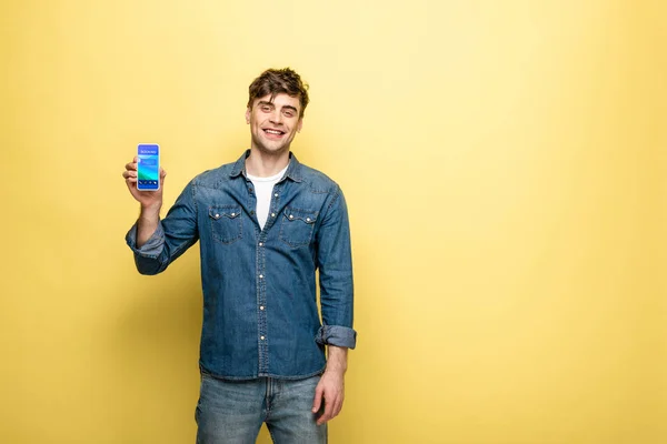 Handsome smiling man in denim clothes holding smartphone with booking app on yellow — Stock Photo